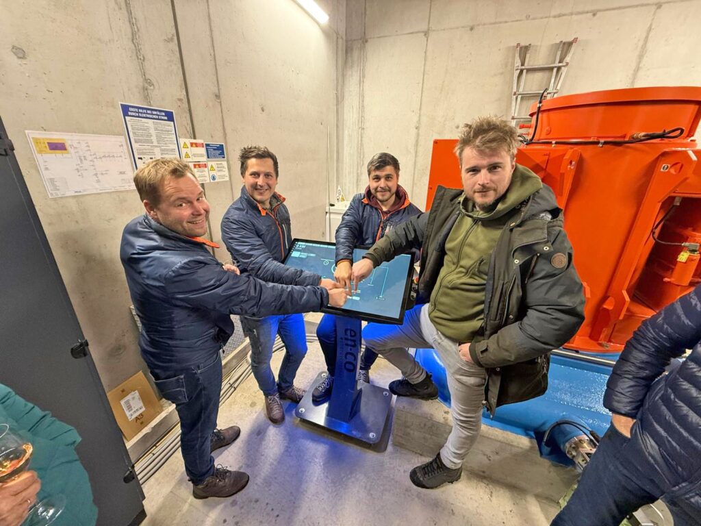 Vier Männer bedienen gemeinsam ein Touchpad-Steuerungssystem in einem Technikraum mit einer großen orangefarbenen Maschine.