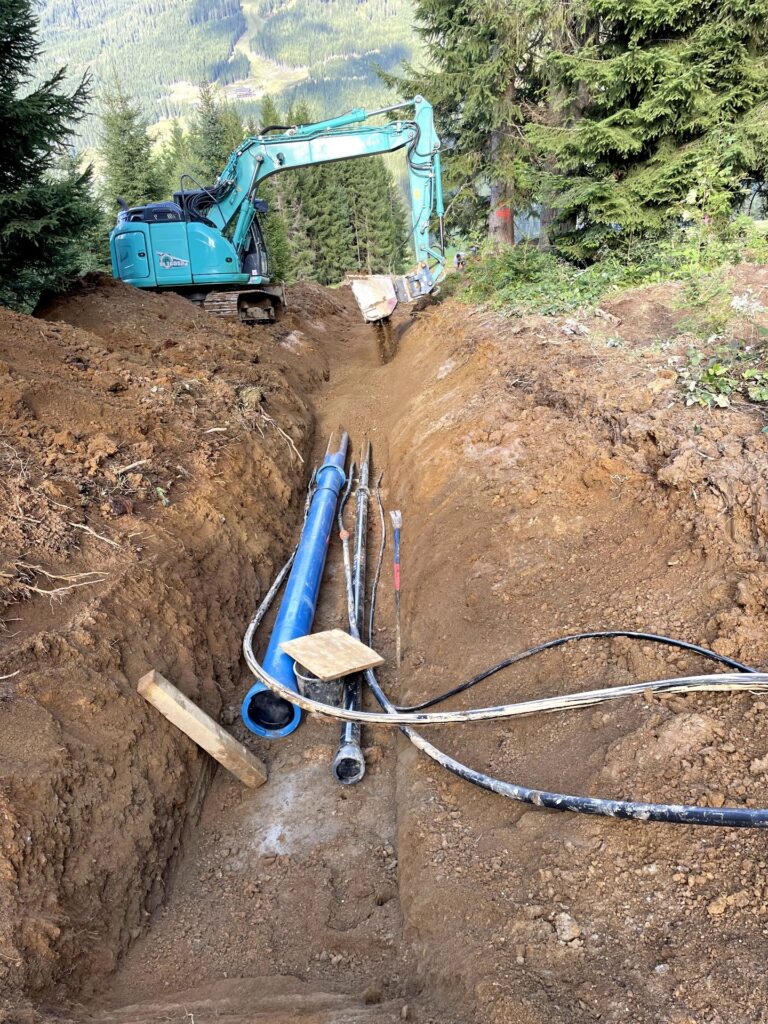Blauer Bagger bei Erdarbeiten im Wald; Wasserrohre und Kabel liegen in einem tiefen Graben für Tiefbauarbeiten am Hang.