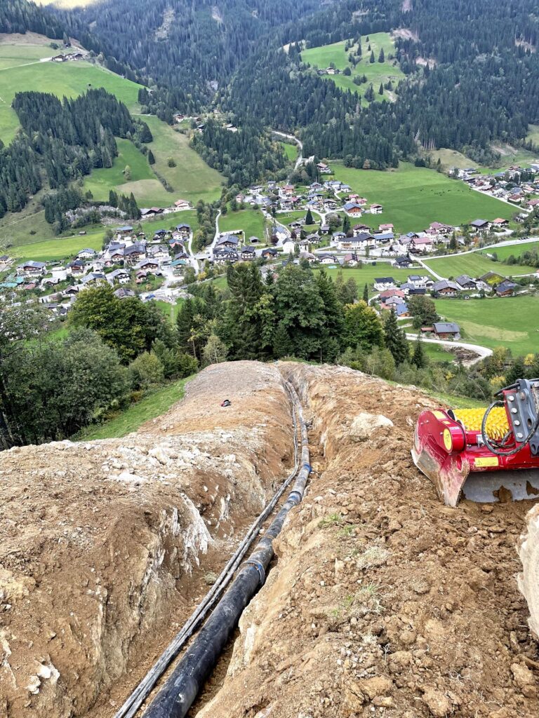 Baugrube am Steilhang mit verlegten schwarzen Rohrleitungen, Blick auf ein Bergdorf in den Alpen und bewaldete Hänge.