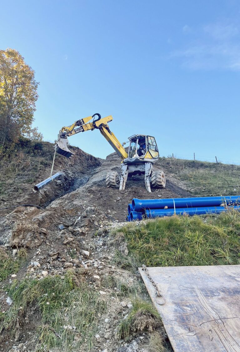 Schreitbagger verlegt Rohre am steilen Hang; Bauarbeiter in Warnweste und blaue Leitungsrohre auf einer Erdbaustelle.