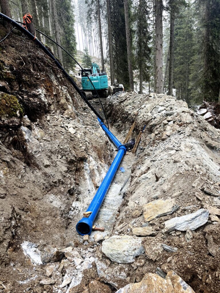 Blaues Gussrohr in einem felsigen Graben im Wald, im Hintergrund ein Kobelco Bagger bei Tiefbauarbeiten am Hang.