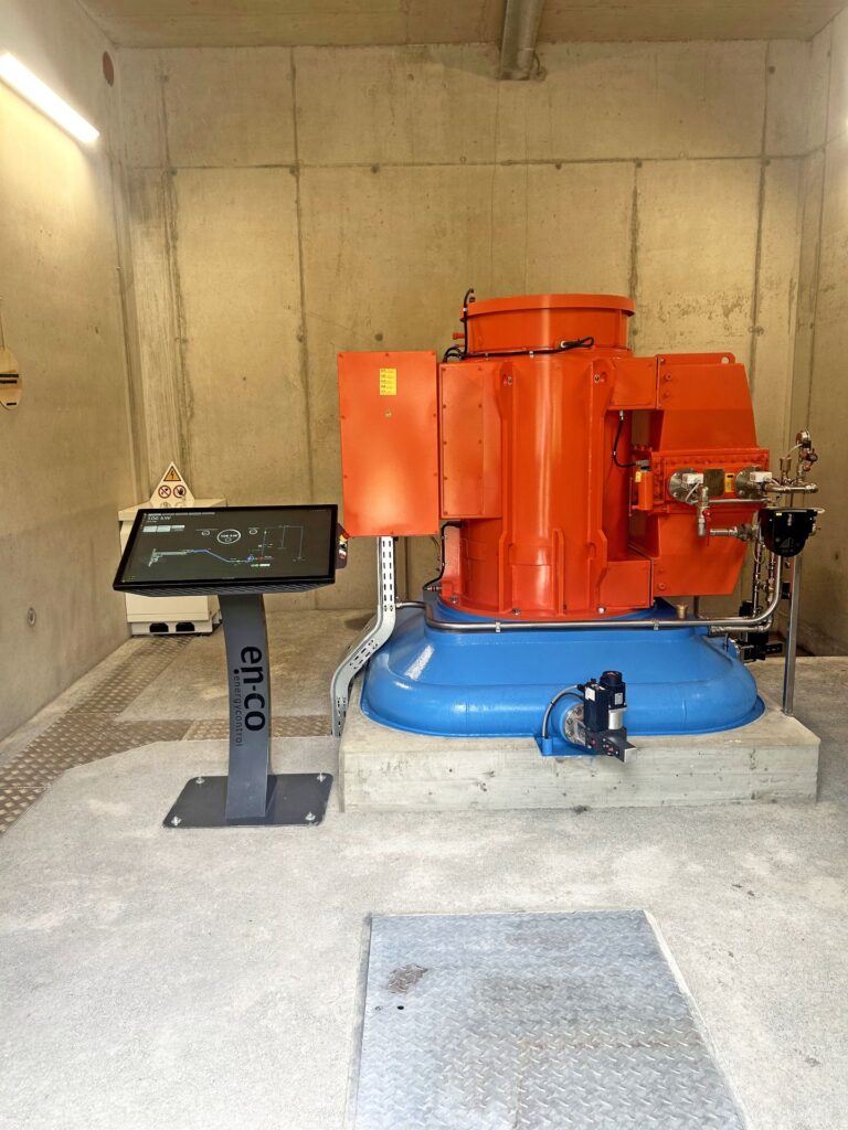 Orangene Wasserturbine auf blauem Sockel mit en-co Energycontrol Bedienterminal in einem Betonraum.