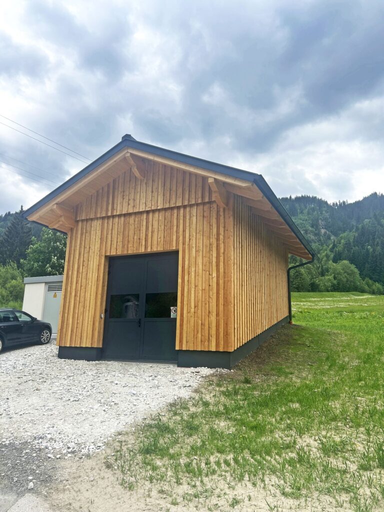 Modernes Holzhaus mit Satteldach, vertikaler Holzfassade und schwarzem Doppeltor vor bewaldeter Gebirgskulisse.
