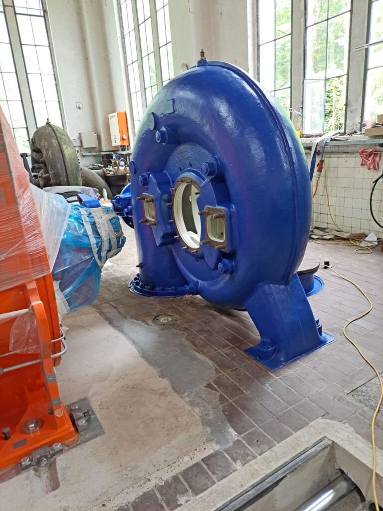 Große blaue Wasserturbine in einer Industriehalle mit hohen Fenstern und Fliesenboden neben einer orangefarbenen Maschine.