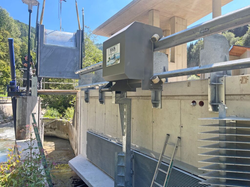 S.K.M. Rechenreinigungsmaschine an einer Wehranlage aus Beton mit Stahlgitter und Kransystem bei einem Wasserkraftwerk.