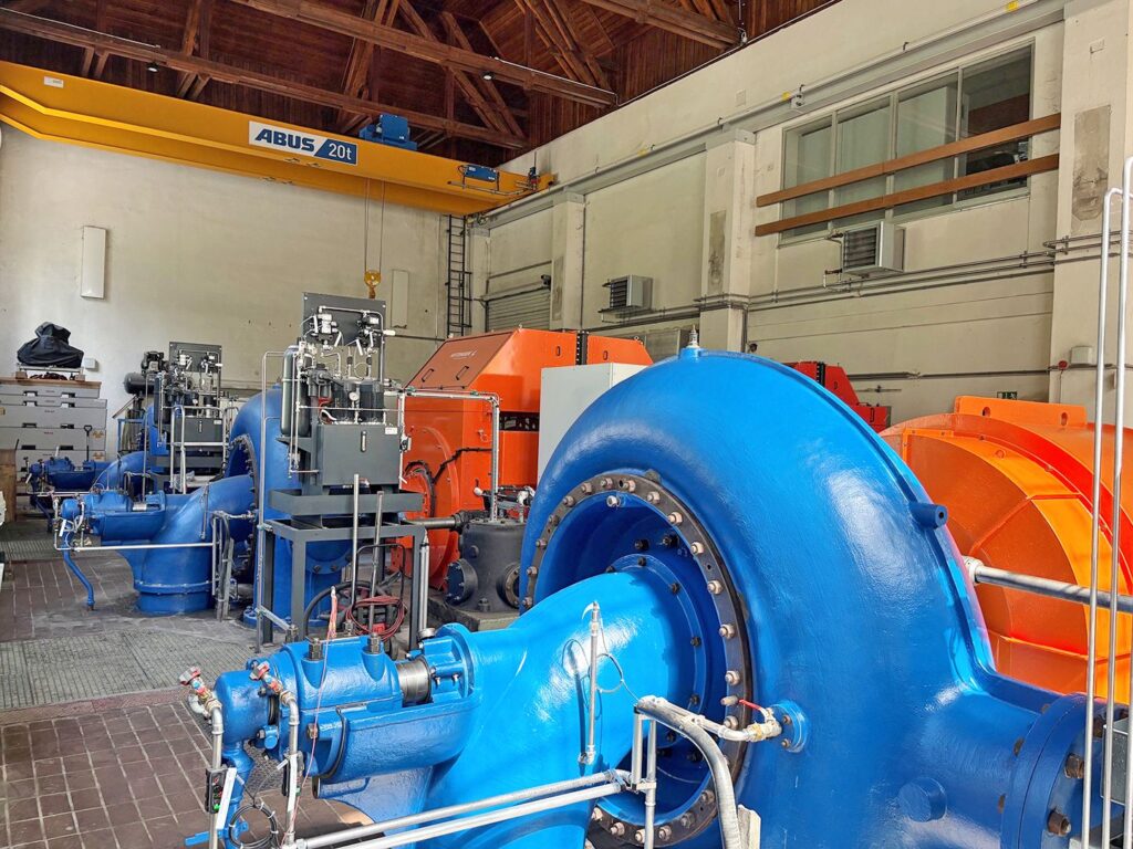 Blaue Turbinen und orangefarbene Generatoren in der Maschinenhalle des KW Erlaufboden mit gelbem Deckenkran.