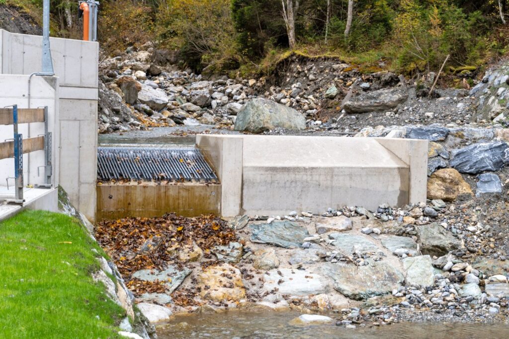 Wasserfassung mit Tiroler Wehr der KW Klaus und Andexer in steinigem Flussbett, Betonstruktur mit Metallrechen im Bergwald.