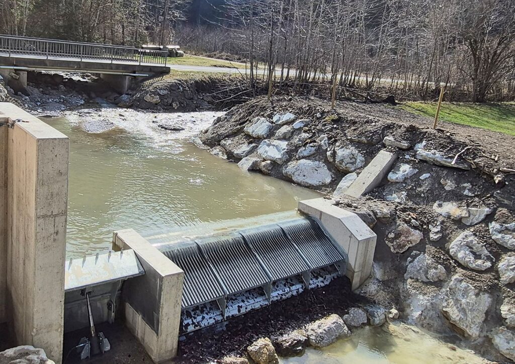 Coanda-Rechen am Kraftwerk Mühlbach mit Wasserüberlauf, Betonmauer und steinerner Uferbefestigung neben einer Brücke.