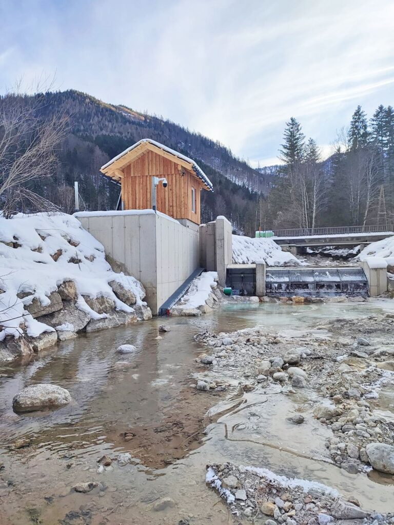 KW Mühlbach Denilpass: Fischaufstiegshilfe und Holzhaus an einem verschneiten Gebirgsbach mit Wehr im Winter.
