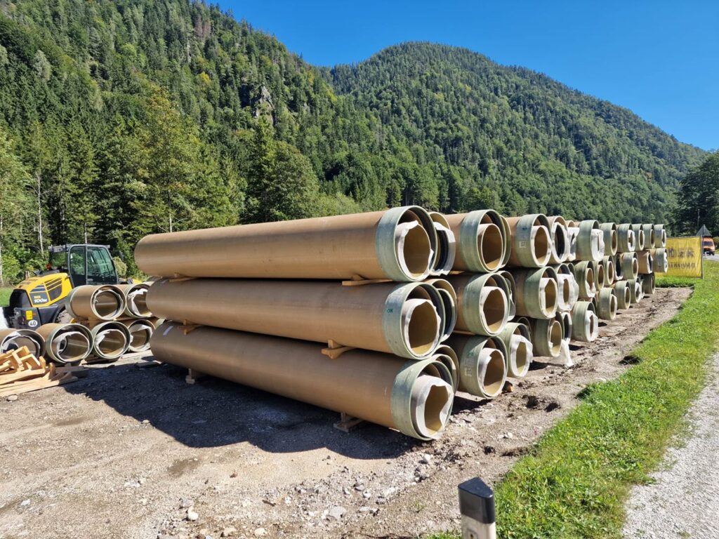 Große GFK-Druckrohre für das KW Mühlbach gestapelt auf einer Baustelle vor bewaldeter Gebirgskulisse und Radlader.