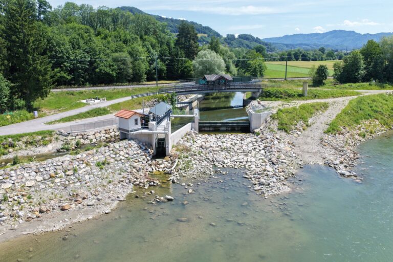 Luftaufnahme vom Kraftwerk Muntigl mit Wehr, Fischaufstiegshilfe und Brücke über den Fluss in grüner Landschaft.