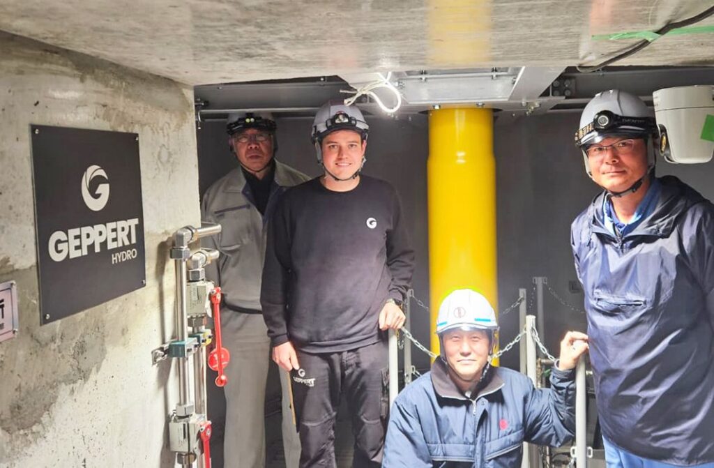 Gruppenfoto im KW Uchiyama: Vier Arbeiter mit Helmen neben Geppert Hydro Logo in einem Wasserkraftwerk.