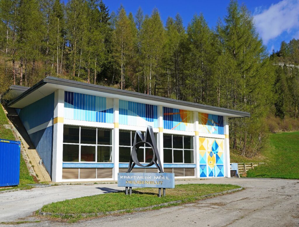 Modernes Gebäude vom Kraftwerk Möll in Heiligenblut vor bewaldetem Hang mit blauer Fassadengestaltung und Skulptur.