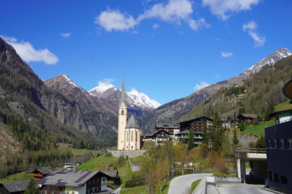 Heiligenblut am Großglockner im Mölltal: Dorfkirche vor schneebedeckten Alpengipfeln unter blauem Himmel in Kärnten.