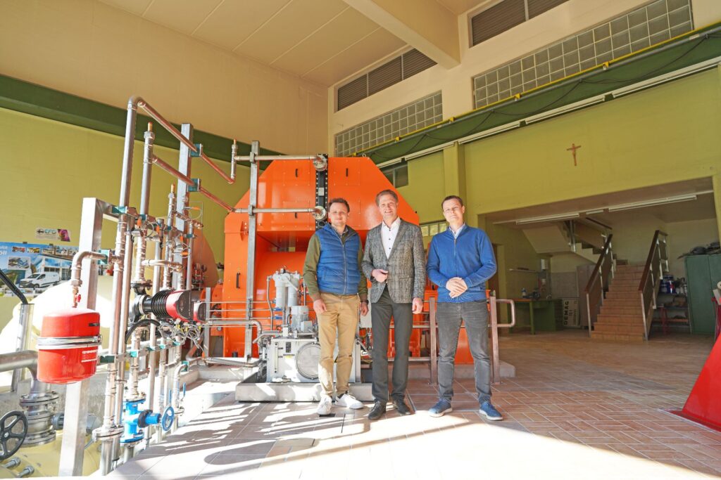 Drei Männer vor einer orangen Turbine im Kraftwerk Möll; industrielle Rohrleitungen und Innenansicht der Maschinenhalle.