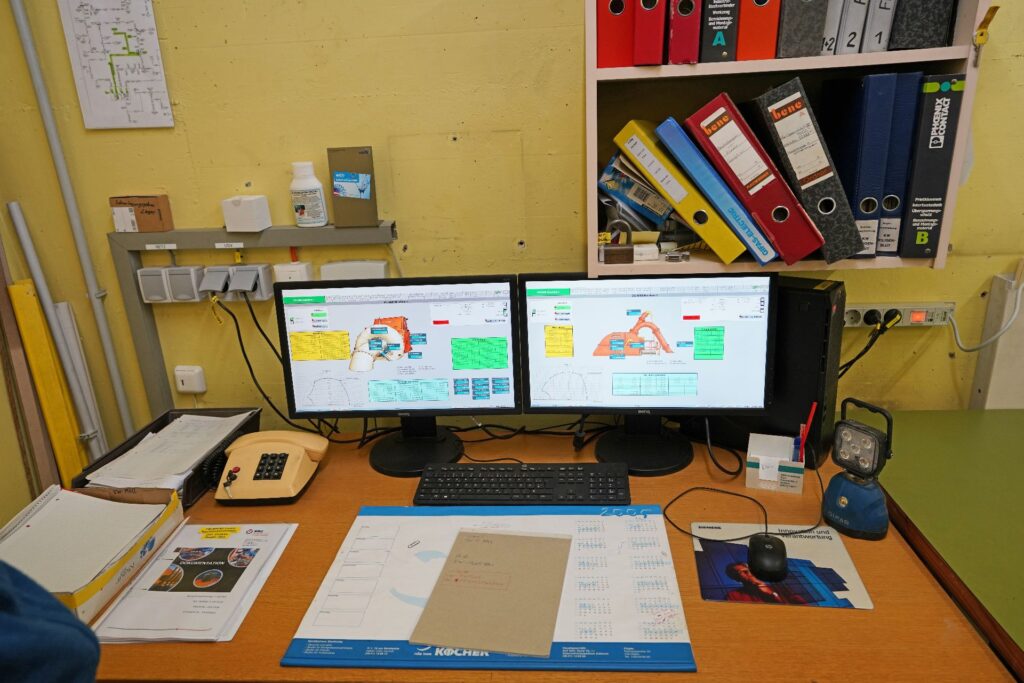 Leitstand Kraftwerk Möll mit zwei Monitoren, technischer Software, Unterlagen und Aktenordnern auf einem Schreibtisch.