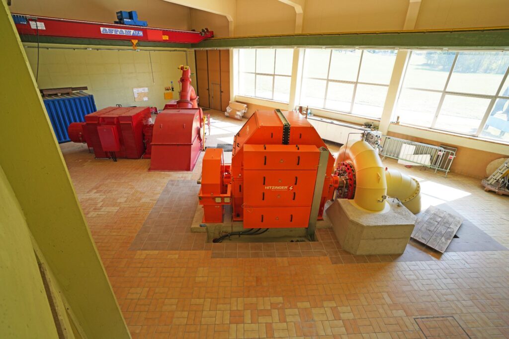 Innenansicht Kraftwerk Möll: Orangefarbener Hitzinger Generator und rote Turbine in einer Industriehalle mit Brückenkran.