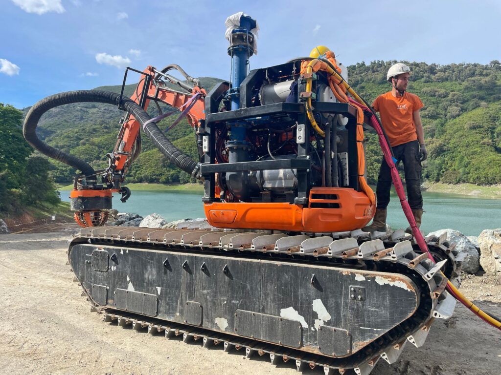Lisie Alesiani Kettenbagger mit Spezial-Brunnenbohrgerät und Arbeiter an einem See vor bewaldeten Bergen.