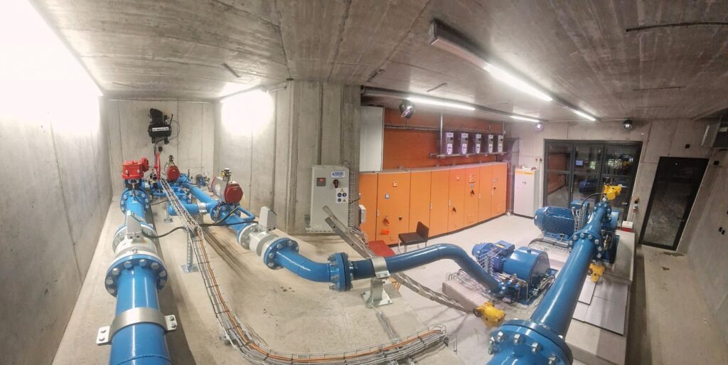 Industrieanlage Fischpass mit blauen Rohrleitungen, Pumpen und orangefarbenen Steuerschränken in Betonraum.