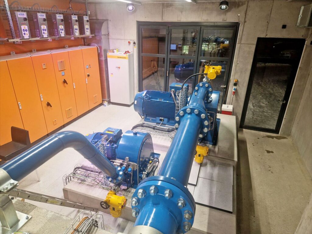 Industrielles Maschinenhaus mit blauen Wasserpumpen, massiven Rohrleitungen und orangefarbenen Schaltschränken in Betonraum.