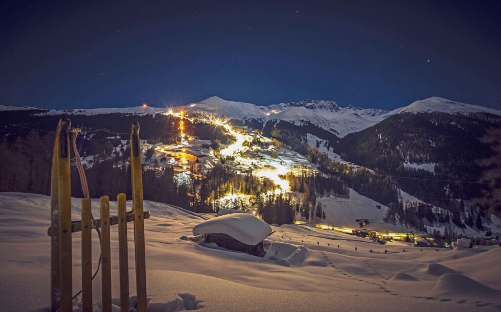 Holzschlitten im Schnee vor beleuchteter Piste zum Nachtschlitteln am Rinerhorn in Davos bei Nacht.