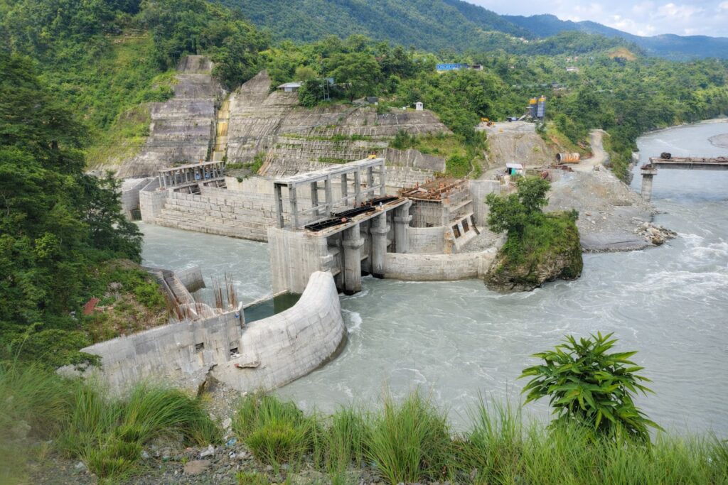 Wasserkraftwerk-Baustelle am Seti Khola Fluss in Nepal mit Betonstaumauer und grünen Berghängen im Hintergrund.