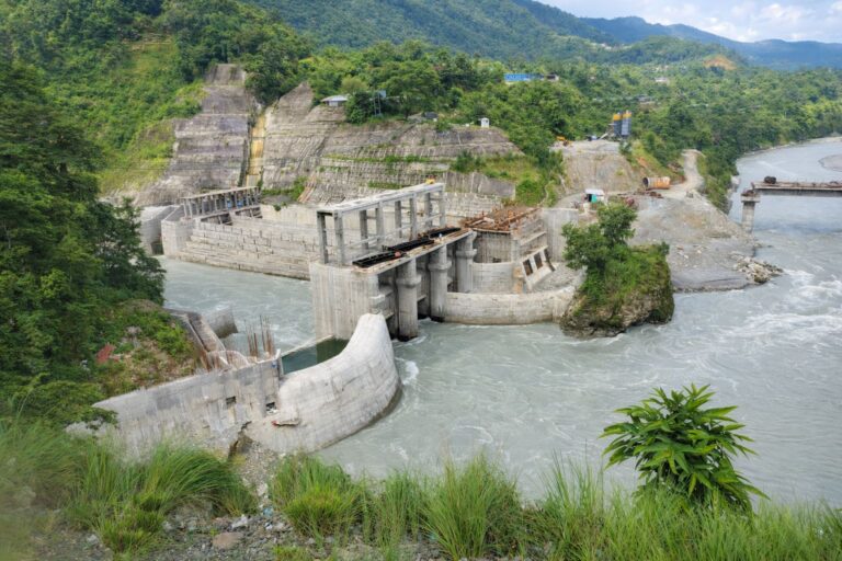 Wasserkraftwerk-Baustelle am Seti Khola Fluss in Nepal mit Betonstaumauer und grünen Berghängen im Hintergrund.