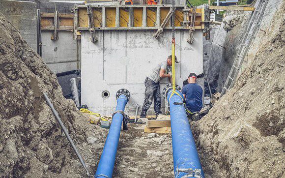 Bauarbeiter montieren einen blauen Rohranschluss an einem Betonbauwerk in einer tiefen Baugrube.