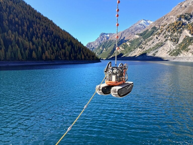 Schwimmender Saugbagger B2 an Seilen über einem blauen Bergsee, umgeben von bewaldeten Hängen und schneebedeckten Gipfeln.