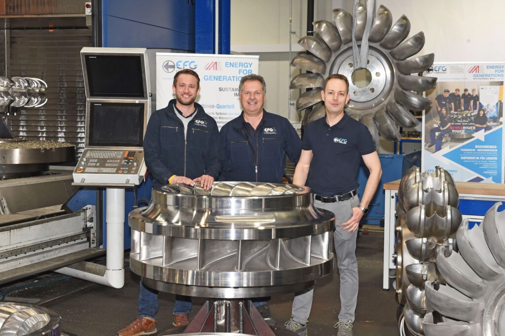 Drei lachende EFG-Mitarbeiter stehen in einer Werkstatt vor einer großen, metallischen Wasserturbine und Steuerungstechnik.