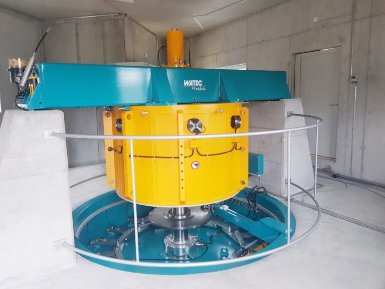 Gelbe Watec Hydro Wasserturbine in einem Betonraum mit blauer Halterung und Metallgeländer zur Energieerzeugung.