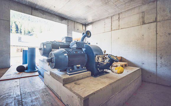 Blaue Wasserturbine bei der Turbinenmontage in einem Betonbau mit großen Rohrleitungen und Blick auf Bäume.