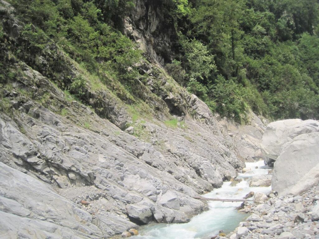 Steile Felswände und grüner Wald am Ufer des Upper Madi Flusses in Nepal mit fließendem Wasser über helle Steine.