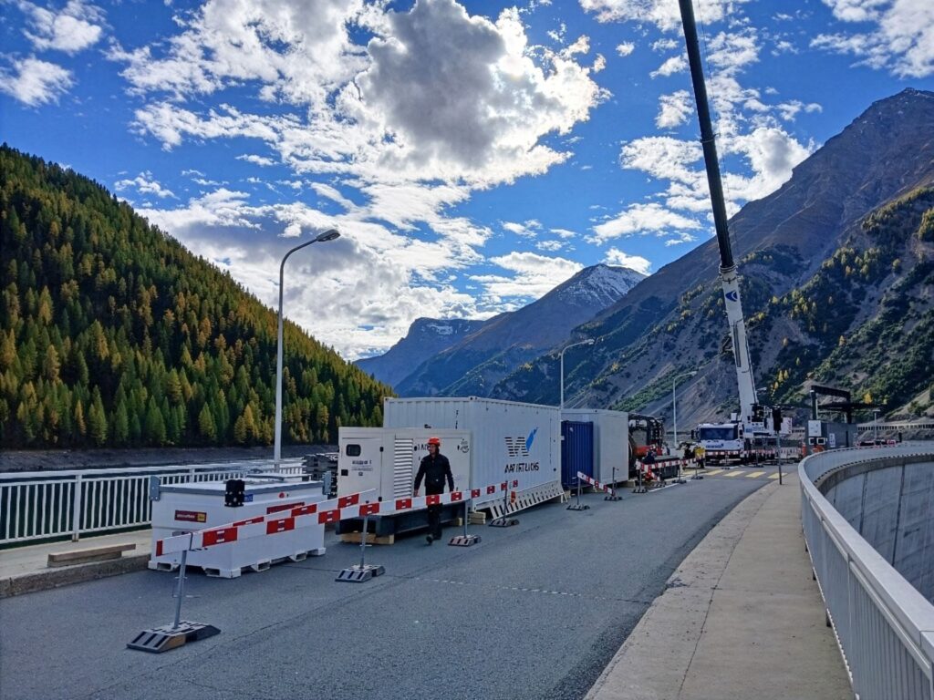 Baustelle an einer Wehrkrone mit Kränen und Containern vor einem alpinen Bergpanorama bei Sonnenschein.