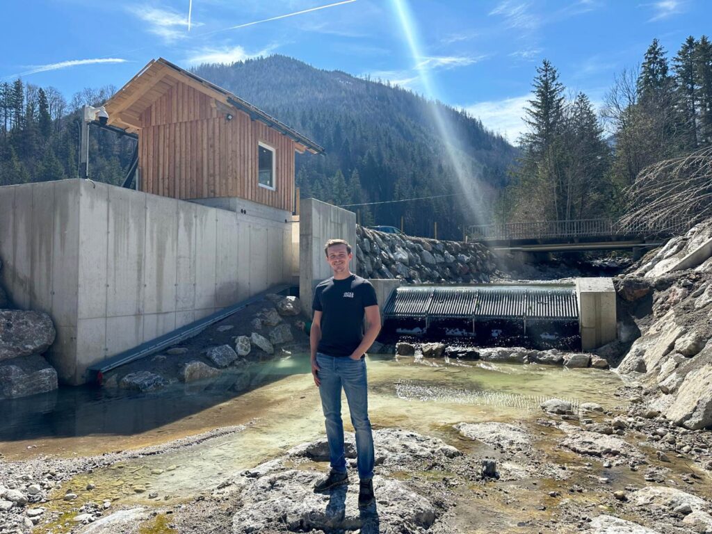 Mann vor modernem Wasserkraftwerk in den Alpen, Holzhaus auf Betonfundament und Rechenanlage vor Gebirgskulisse.