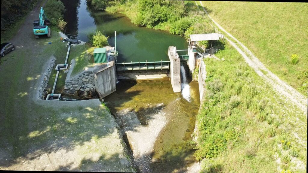 Luftaufnahme eines Wehrs am Fluss mit Fischtreppe, Wasserfall und blauem Bagger der Firma Erdbau am Ufer.