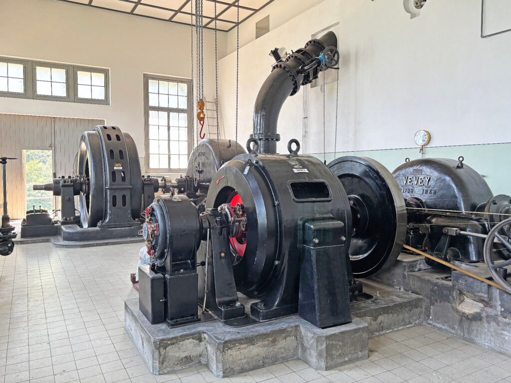 Historische schwarze Vevey Turbinen und Generatoren in einem hellen Maschinenraum mit großen Fenstern und Betonsockeln.