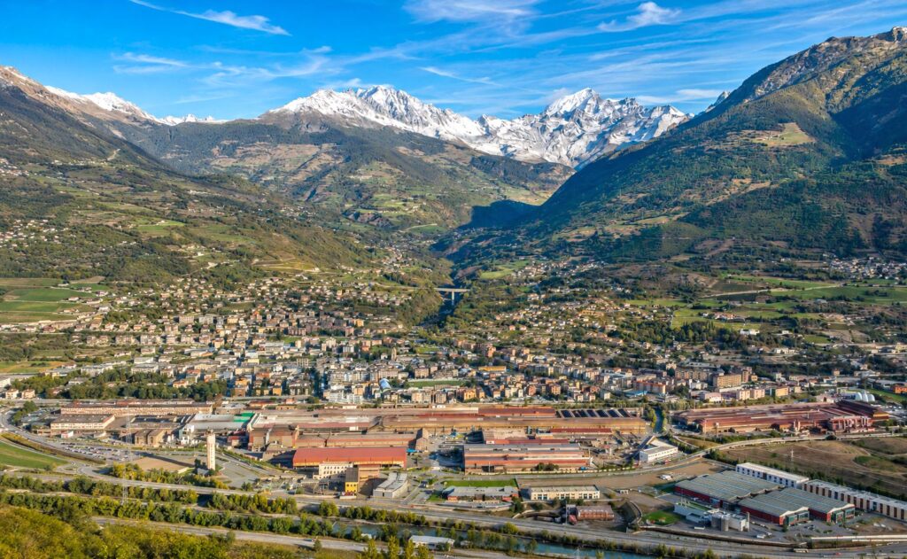Panoramablick auf Aosta im Aostatal mit Industriegebäuden, Wohnvierteln und schneebedeckten Alpengipfeln unter blauem Himmel.