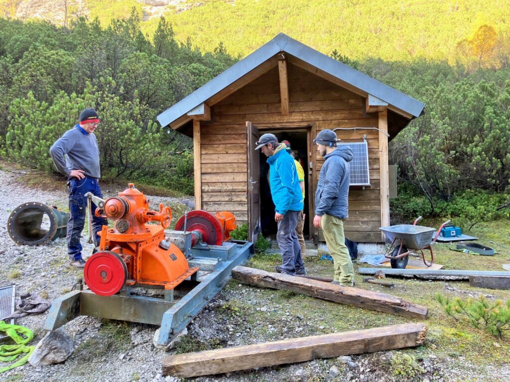 Drei Männer beim Ausbau einer technischen Anlage vor einer Holzhütte in den Bergen mit orangefarbener Maschine auf Schienen.