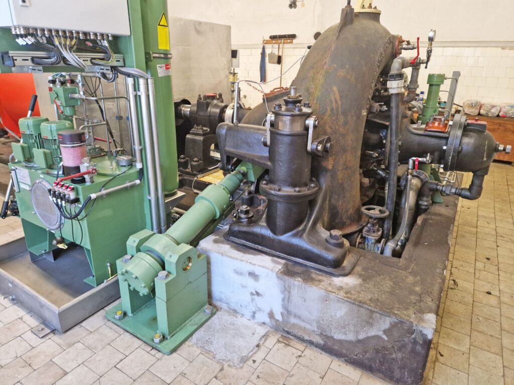 Industrielle Wasserturbine mit gusseisernem Gehäuse, grüner Steuereinheit und Rohrleitungen in einem Maschinenraum.