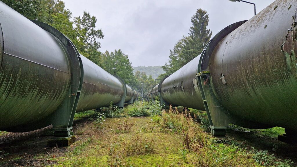 Zwei riesige, parallel verlaufende Rohre aus Metall in einer bewaldeten Landschaft unter grauem Himmel.