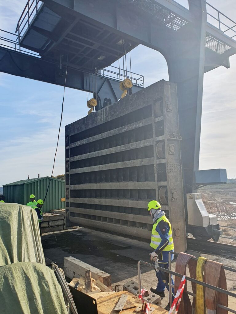 Arbeiter mit Sicherheitskleidung heben ein massives Stahlschütz an einer Krananlage auf einer Industrie-Baustelle.