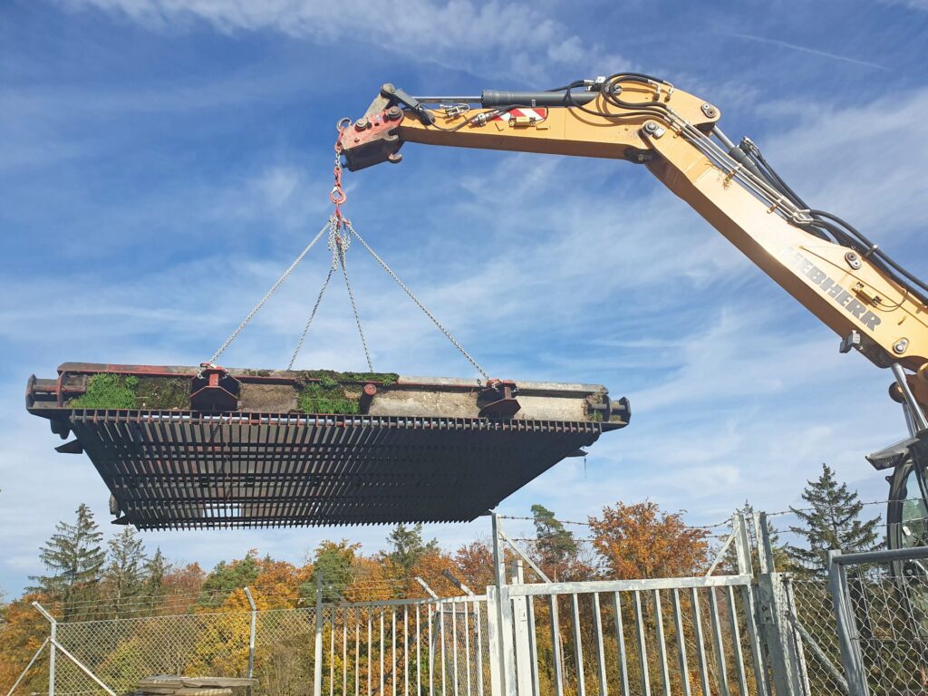 Gelber Liebherr Bagger hebt ein schweres Metallgitter an Ketten vor blauem Himmel und herbstlichen Bäumen.