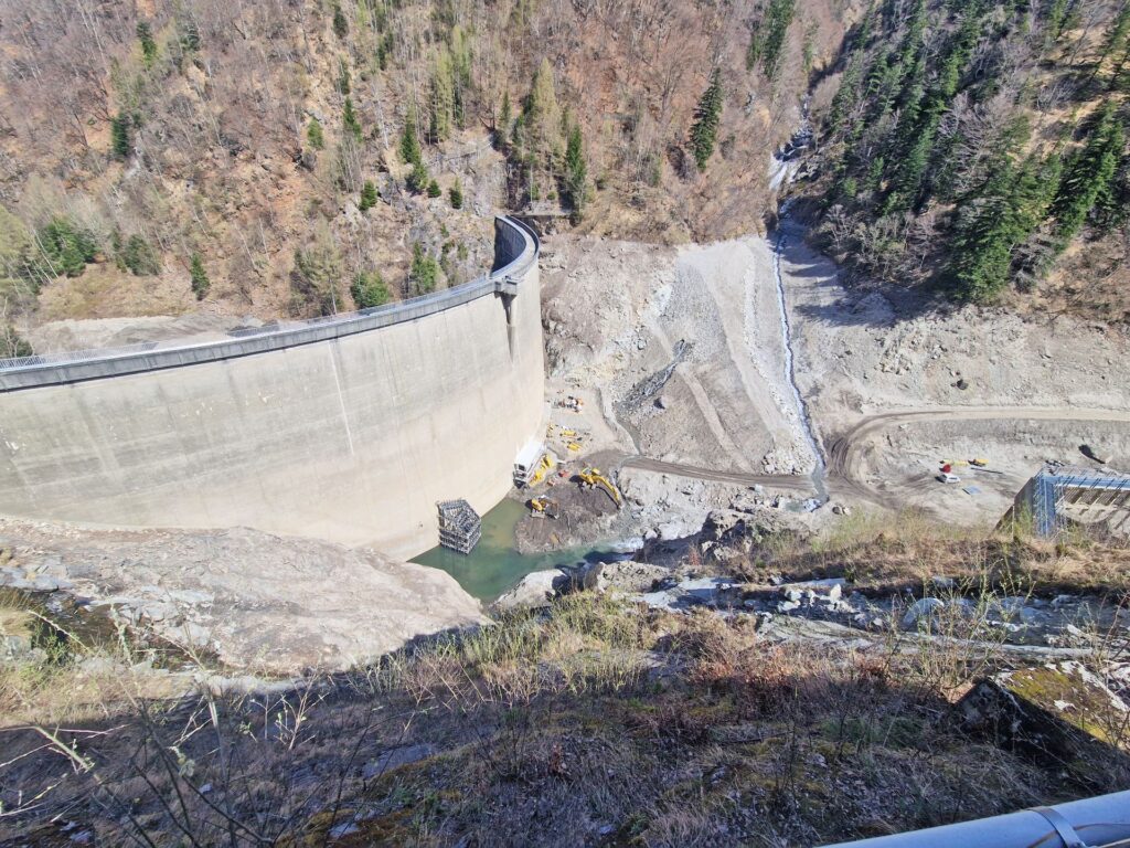 Gekrümmte Staumauer aus Beton in einer bewaldeten Gebirgsschlucht mit Baustelle und gelben Baggern am Wandfuß.