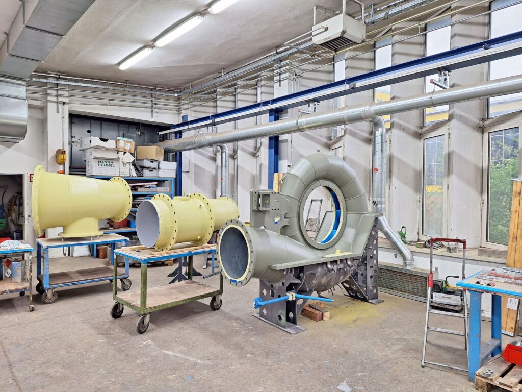 Industrielle Fertigungshalle mit großen, beigefarbenen Rohrleitungselementen und einem massiven Turbinengehäuse auf Ständern.