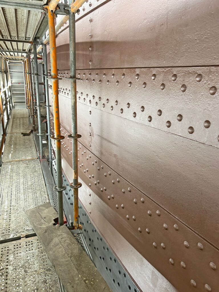 Metallgerüst vor einer braun gestrichenen, genieteten Stahlwand einer Brücke oder eines Schiffes bei Sanierungsarbeiten.