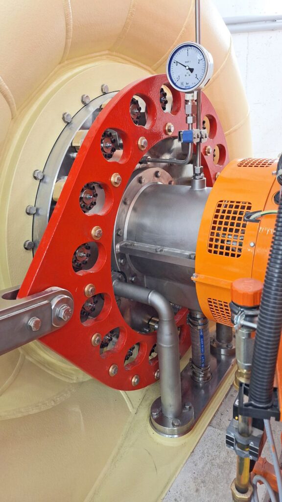 Industrielle Wasserturbine mit rotem Stellring, Manometer und orangefarbenem Generatorgehäuse in einer Kraftwerksanlage.