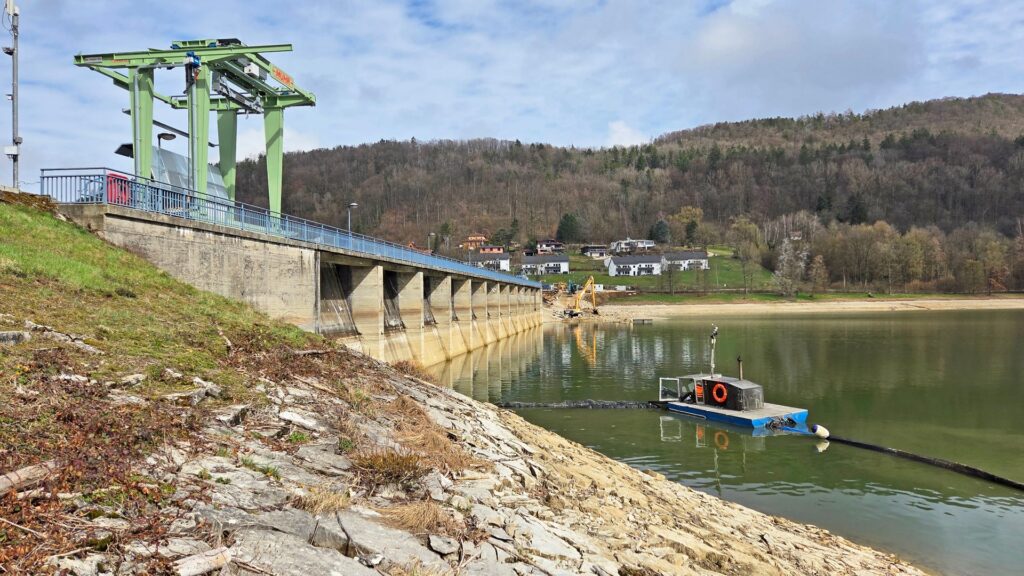 Beton-Staumauer mit grünem Portalkran und Arbeitsboot auf einem Stausee vor bewaldetem Hang und Häusern.