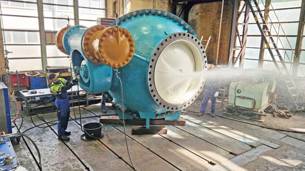 Industrielle Hochdruckreinigung eines großen, blau-gelben Kugelventils in einer Werkshalle durch einen Arbeiter.