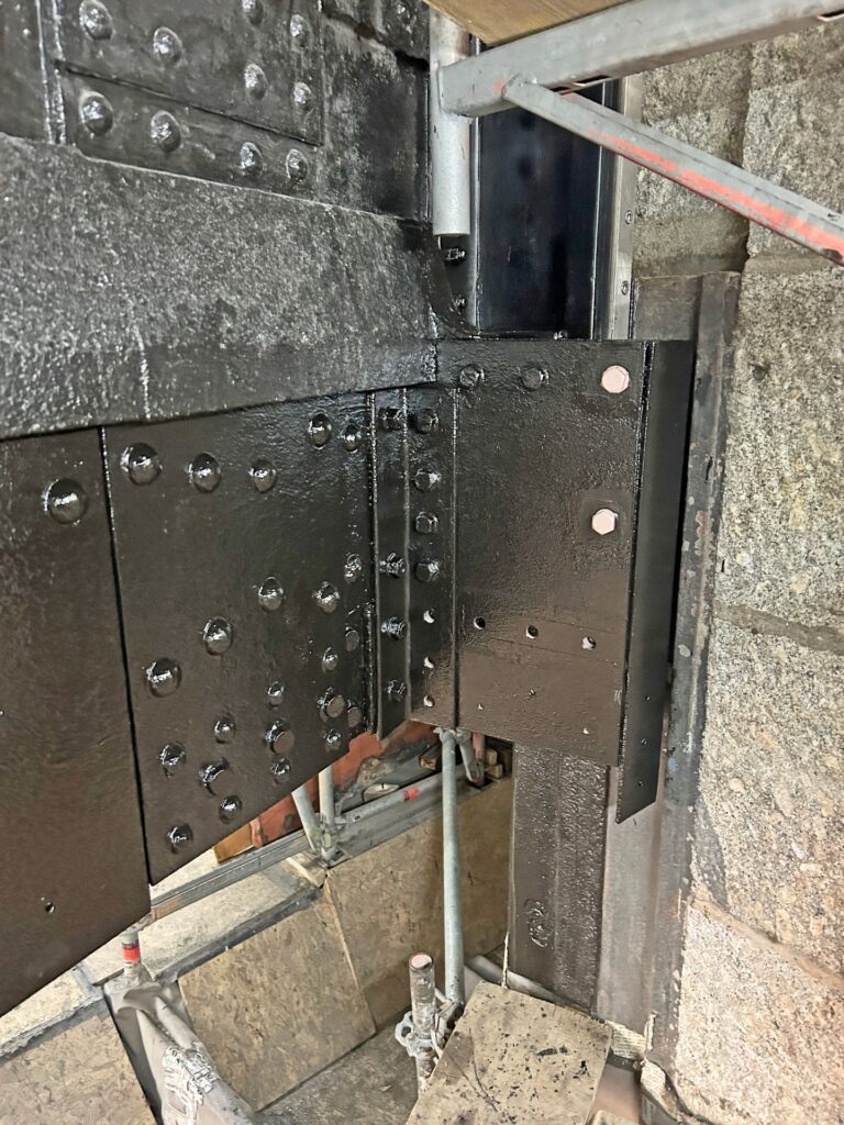 Schwarz lackierte Stahlträger mit Nieten an einer Steinfassade auf einer Baustelle mit Gerüstteilen.